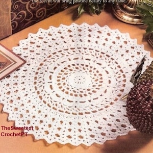 May include: White crochet doily with a delicate lace pattern. The doily is round and has a center circle with a smaller, intricate design. The doily is on a brown surface.