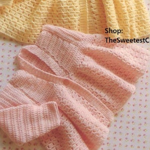 May include: A handmade, light yellow crochet baby cardigan with button closures and a flared skirt. A matching pink crochet baby cardigan is folded below. The text "Shop: TheSweetestCrochetPT" is visible.