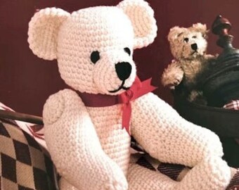 Può includere: Un orsacchiotto all'uncinetto color crema con occhi e naso neri, che indossa un nastro rosso annodato a fiocco intorno al collo. Il testo TheSweetestCrochetPT è visibile sul lato inferiore destro dell'orso. L'orso ha una superficie in filato testurizzato.