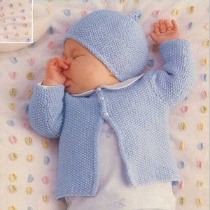 May include: A baby wearing a light blue knitted cardigan, hat and booties. The baby is lying on a white and pink patterned blanket.