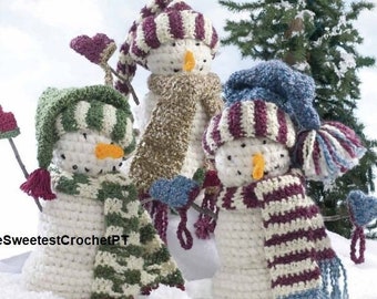 May include: Three crocheted snowmen figures with white bodies and orange noses. Each wears a unique hat and scarf set in green, maroon, or blue stripes. They hold small crocheted heart-shaped ornaments on sticks. The text TheSweetestCrochetPT is visible.
