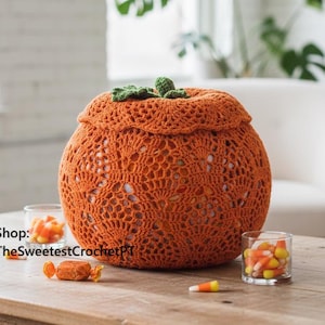May include: An orange crochet pumpkin with a green stem and leaves sits on a wooden table. The pumpkin has an openwork design. Candy corn and wrapped candies are scattered on the table. The text "Shop: TheSweetestCrochetPT" is visible.