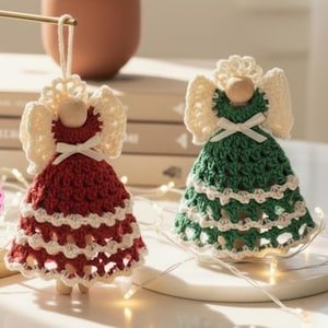 May include: Two handmade crochet angel ornaments. One is red and cream, the other is green and cream. Both have wooden heads, cream-colored wings, and small white bows. They are displayed on a white surface with fairy lights.