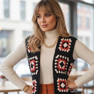 May include: A long, open-front crochet vest in black with red and white square patterns. The vest is worn over a cream-colored turtleneck and rust-colored pants. A gold chain necklace and bracelets complete the look. The shop name is visible in the image.