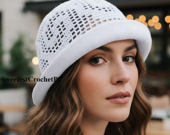 May include: A white crochet bucket hat featuring a geometric openwork pattern and a solid brim. The text TheSweetestCrochetPL is visible on the left side of the hat. The hat is constructed from yarn with a visible texture and a rounded crown shape.