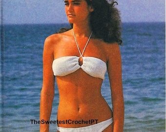 May include: A white two-piece crochet bikini set featuring a bandeau-style top with a central ring detail and a matching bottom. The text TheSweetestCrochetPT is visible in the center. A bright yellow towel is held in one hand.