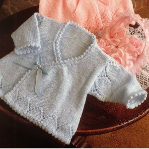 Op de afbeelding: Twee gebreide babytruien, één roze en één lichtblauw, met een kantpatroon en een strikdetail.