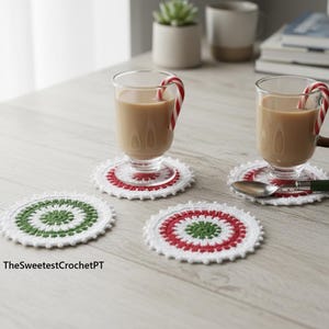 May include: A cozy scene featuring two clear glass mugs with brown handles, each holding a warm beverage and a candy cane. The mugs rest on white, red, and green crocheted coasters. Additional coasters are visible, along with a spoon.