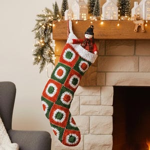Puede incluir: Un calcetín navideño de ganchillo con un patrón de cuadrados de la abuela en rojo, blanco y verde, colgado de una repisa de chimenea. Una pequeña muñeca con sombrero negro y camisa de cuadros rojos se asoma por la abertura. El texto Shop: TheSweetestCrochetPT es visible.