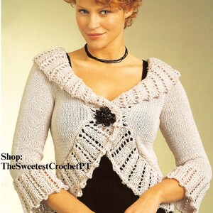 Könnte beinhalten: Hellbeige gestrickte Bolero-Jacke mit langen Ärmeln, Rüschenkragen und -bündchen. Die Jacke hat eine dekorative schwarze Verzierung in der Mitte und einen V-Ausschnitt. Der Text "Shop: TheSweetestCrochetPT" ist sichtbar.