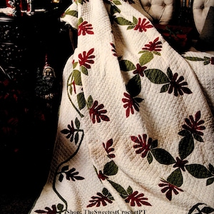 Puede incluir: Una colcha blanca con un patrón repetitivo de motivos florales rojo oscuro y aplicaciones de hojas verdes. La tela tiene una textura acolchada en toda su superficie y el texto Shop: TheSweetestCrochetPT aparece en la parte inferior central.
