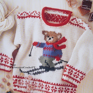 May include: Cream-colored knitted sweater with a red and white pattern on the cuffs and hem. The sweater features a bear skiing design with a red scarf. The neckline is trimmed in red. The text "Shop: TheSweetestCrochetPT" is visible.