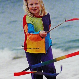 May include: A young girl wearing a colourful crocheted sweater and purple trousers plays with a rainbow ribbon on a beach.
