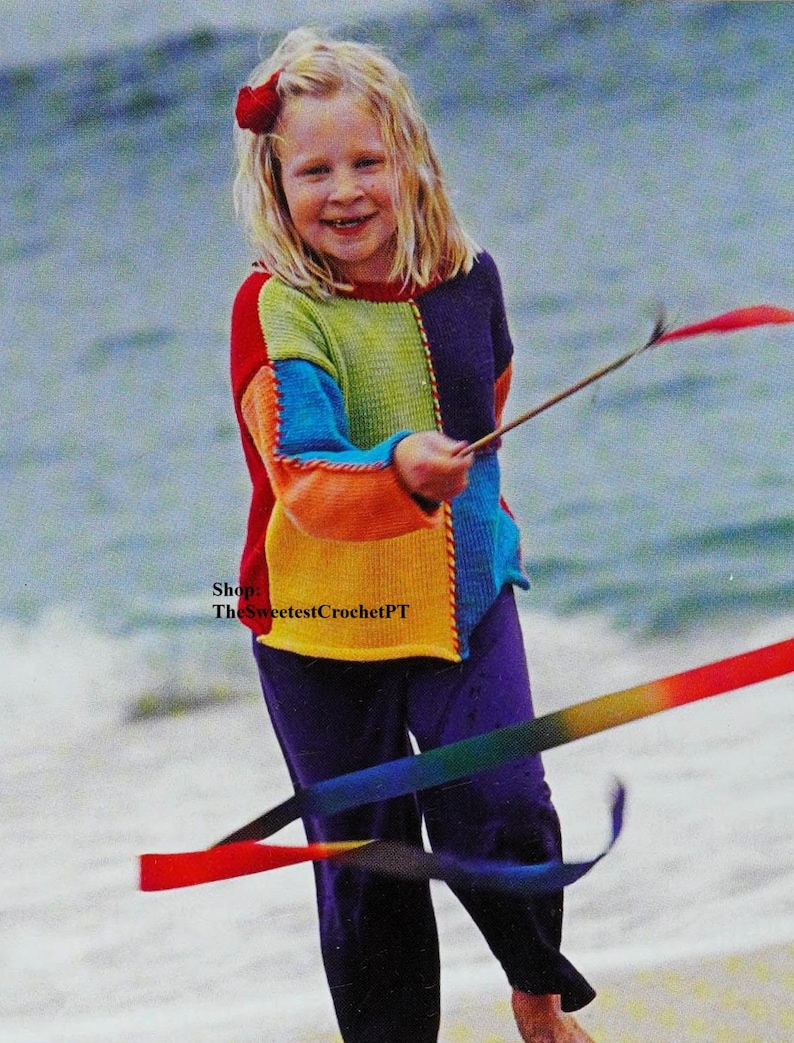 May include: A young girl wearing a colourful crocheted sweater and purple trousers plays with a rainbow ribbon on a beach.