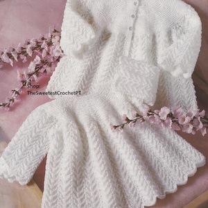 Pode incluir: Conjunto de bebê de malha branca de duas peças. O conjunto inclui um cardigan de manga comprida com três botões e um vestido com bainha ondulada. As peças de vestuário têm um padrão de chevron texturizado. Flores cor-de-rosa no fundo. Shop: TheSweetestCrochetPT.