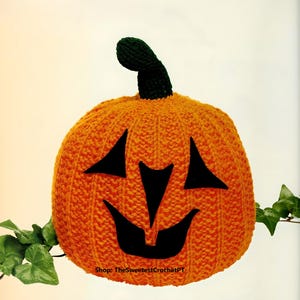 Op de afbeelding: Een feloranje gehaakte pompoen met een donkergroene steel en zwarte vilten gezichtskenmerken, waaronder twee driehoekige ogen en een brede, grillige mond. De tekst Shop: TheSweetestCrochetPT is in het zwart onderaan de pompoen gedrukt.