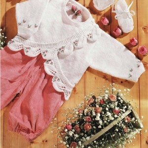 May include: A white knitted baby outfit with pink corduroy pants, a sweater with a scalloped collar, and matching booties. The outfit is decorated with small pink floral embroidery. A wooden box of flowers and scattered roses complete the scene.