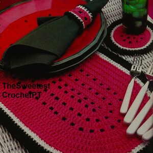 May include: A crocheted watermelon placemat with black and white trim. The placemat is pink and has a white and black crocheted napkin ring on a red plate.