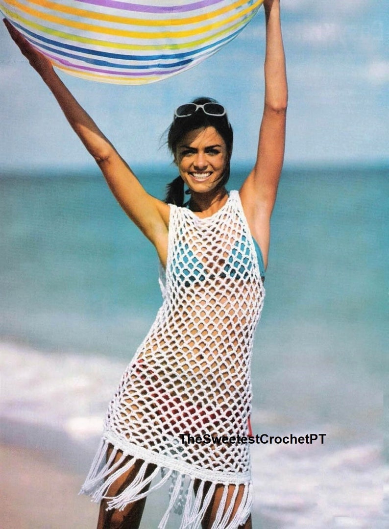 May include: A white sleeveless crochet cover-up dress featuring an open diamond-shaped mesh pattern and a fringed hemline. The garment is worn over a blue bikini top and patterned bikini bottoms. The text TheSweetestCrochetPT is visible at the bottom.