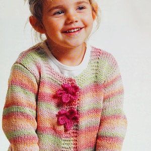 May include: A young girl wearing a striped cardigan with pink flower embellishments. The cardigan is made of a soft, colorful yarn and has a relaxed fit. The girl is smiling and looking to the side.