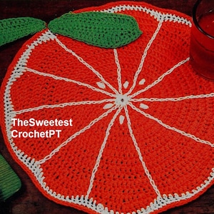 May include: Crocheted orange slice coaster in red and white with a green leaf. The coaster is on a wooden surface with a glass of juice and oranges.