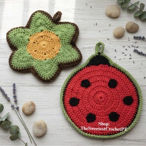Op de afbeelding: Twee handgemaakte gehaakte pannenlappen. Eén is gevormd als een groen en bruin bloem met een gele kern. De andere is een rode lieveheersbeestje met zwarte stippen en een groene rand. Beide hebben een lus om op te hangen.
