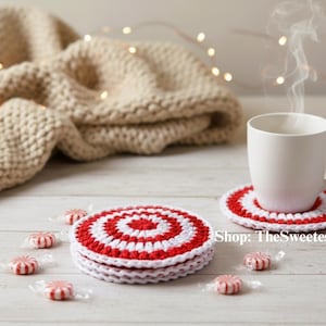 May include: A stack of red and white crocheted coasters with a matching coaster under a white mug. Several wrapped peppermint sweets are scattered around the coasters. A beige knitted blanket is in the background.