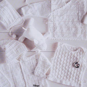 May include: A set of white crocheted baby clothes, including a hooded jacket, a cardigan, a vest, and a hat. The clothes are laid out on a white wooden surface. Shop: TheSweetestCrochetPT
