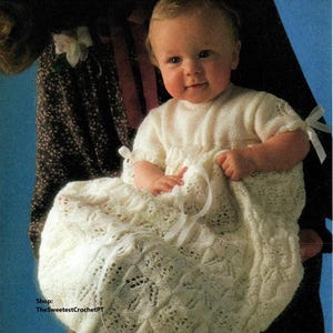 May include: A white crocheted baby dress with a delicate lace pattern and satin ribbon ties. The dress is being held by a person, and the baby is wearing the dress.