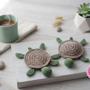 Puede incluir: Dos posavasos de tortuga hechos a mano con ganchillo, con aletas y cabezas verdes y caparazones marrones. Una taza verde claro con una bebida caliente se encuentra sobre una superficie de madera blanca, junto con una pequeña suculenta.