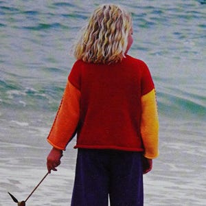 May include: A young person wearing a red, orange and yellow jumper with long sleeves and blue trousers. They are standing on a beach with their back to the camera and holding a stick with a colourful ribbon attached to it.