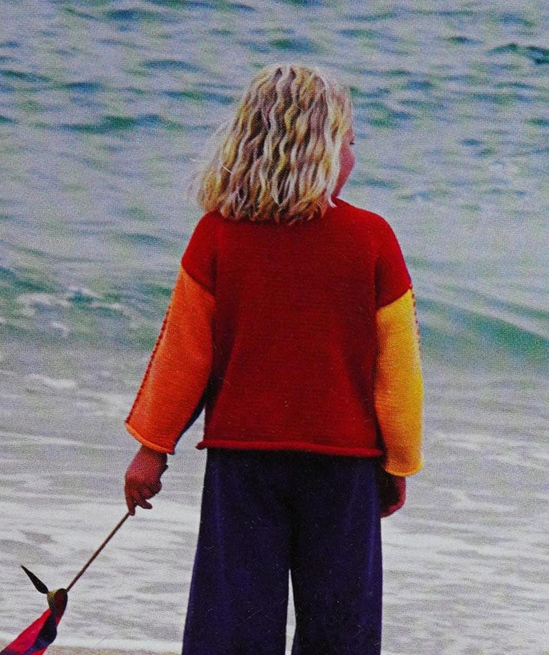 May include: A young person wearing a red, orange and yellow jumper with long sleeves and blue trousers. They are standing on a beach with their back to the camera and holding a stick with a colourful ribbon attached to it.