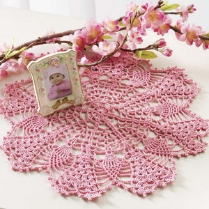 May include: A round, pink, intricate crochet doily with a scalloped edge. A small, rectangular, cream-colored picture frame with floral accents and a gold bow detail sits on the doily, displaying a photograph of a baby in a pink outfit.