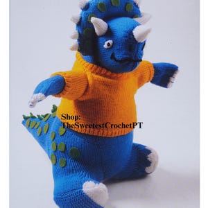 May include: A blue knitted triceratops dinosaur wearing an orange knitted jumper. The dinosaur has green spots on its back and tail. Shop: TheSweetestCrochetPT