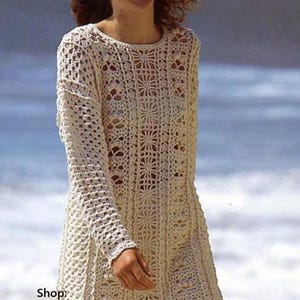 May include: Cream-colored crochet tunic with long sleeves and a floral pattern. The tunic has a round neckline and a loose fit. The text "Shop: TheSweetestCrochetPT" is visible. This handmade garment is perfect for beachwear or a casual summer look.