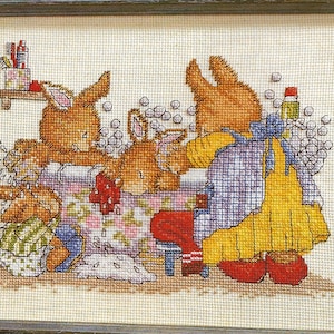 May include: A framed cross-stitch embroidery depicting three brown rabbits in a bathroom setting. One rabbit wears a yellow dress and blue apron, washing another rabbit in a floral bathtub filled with bubbles. A third small rabbit sits nearby.