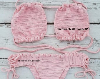 Pode incluir: Um conjunto de biquíni de crochê rosa claro com um top estilo frente única com bordas franzidas e uma parte de baixo combinando com laços laterais. O texto TheSweetestCrochetPT é visível em ambas as peças deste traje de banho de duas peças.