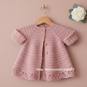 Puede incluir: Un cárdigan de bebé de ganchillo rosa claro, de manga corta, con botones de madera. El cárdigan está colgado de una percha de madera sobre un fondo rosa. El dobladillo inferior presenta un diseño calado decorativo. Una rosa blanca con hojas verdes está en la esquina superior derecha.