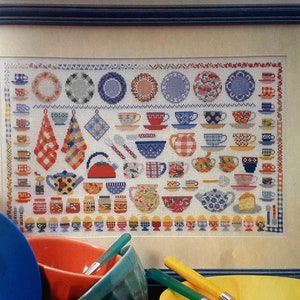 May include: A framed cross-stitch embroidery depicting a variety of kitchenware, including plates, cups, teapots, and other kitchen items. The embroidery is done in a variety of colors, including red, blue, yellow, and green.