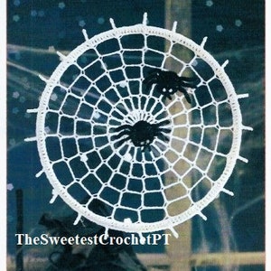 May include: White crocheted spider web with two black spiders, one on the outside and one in the center. The web is round and has a white border. The text "TheSweetestCrochetPT" is visible at the bottom of the image.