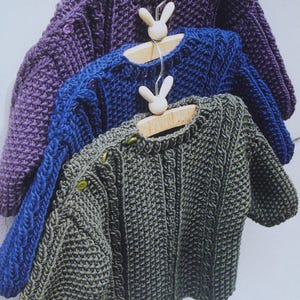 Pode incluir: Três suéteres de crochê em roxo, azul e verde pendurados em cabides de madeira com ganchos em forma de coelho. Os suéteres são todos do mesmo estilo com um padrão de tricô de cabo e fechos de botão. O nome da loja "TheSweetestCrochetPT" está escrito no canto inferior direito.