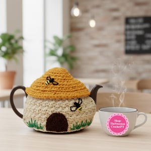 May include: A whimsical teapot cozy shaped like a beehive, with a yellow roof and cream-colored walls. Two embroidered bees and green grass accents add detail. A teacup with steam rising sits beside the teapot. Shop: TheSweetestCrochetPT.