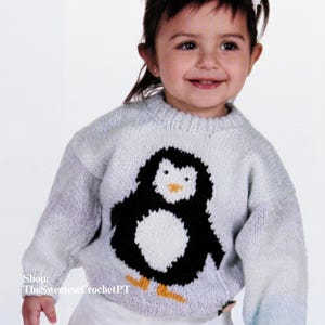 May include: A light gray knitted sweater featuring a large black and white penguin design with an orange beak and yellow feet. The sweater has long sleeves and a round neckline. The text "Shop: TheSweetestCrochetPT" is visible.
