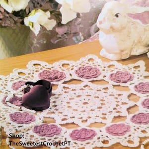 Könnte beinhalten: Eine weiße Häkeldeckchen mit einem zarten Blumenmuster und einer rosa Mitte. Die Deckchen ist mit einer Perlenkette und einer kleinen, dunkelvioletten Samtbox in Herzform verziert. Der Text "Shop: TheSweetestCrochetPT" ist in der linken unteren Ecke des Bildes sichtbar.