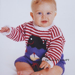Könnte beinhalten: Ein Baby trägt einen rot-weiß gestreiften Strickpullover mit einer blauen Wal-Applikation. Der Pullover hat einen weißen Kragen und lange Ärmel. Das Baby trägt auch blaue Strickhosen mit einer Segelboot-Applikation.