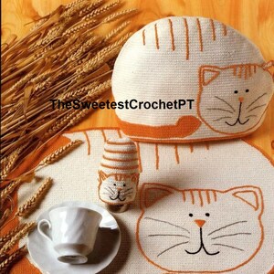 Könnte beinhalten: Eine gehäkelte Teekanne in Katzenform, eine gehäkelte Tasse in Katzenform und ein gehäkeltes Katzen-Platzset. Die Artikel sind alle weiß mit orangefarbenen Akzenten und haben gestickte Katzenköpfe.
