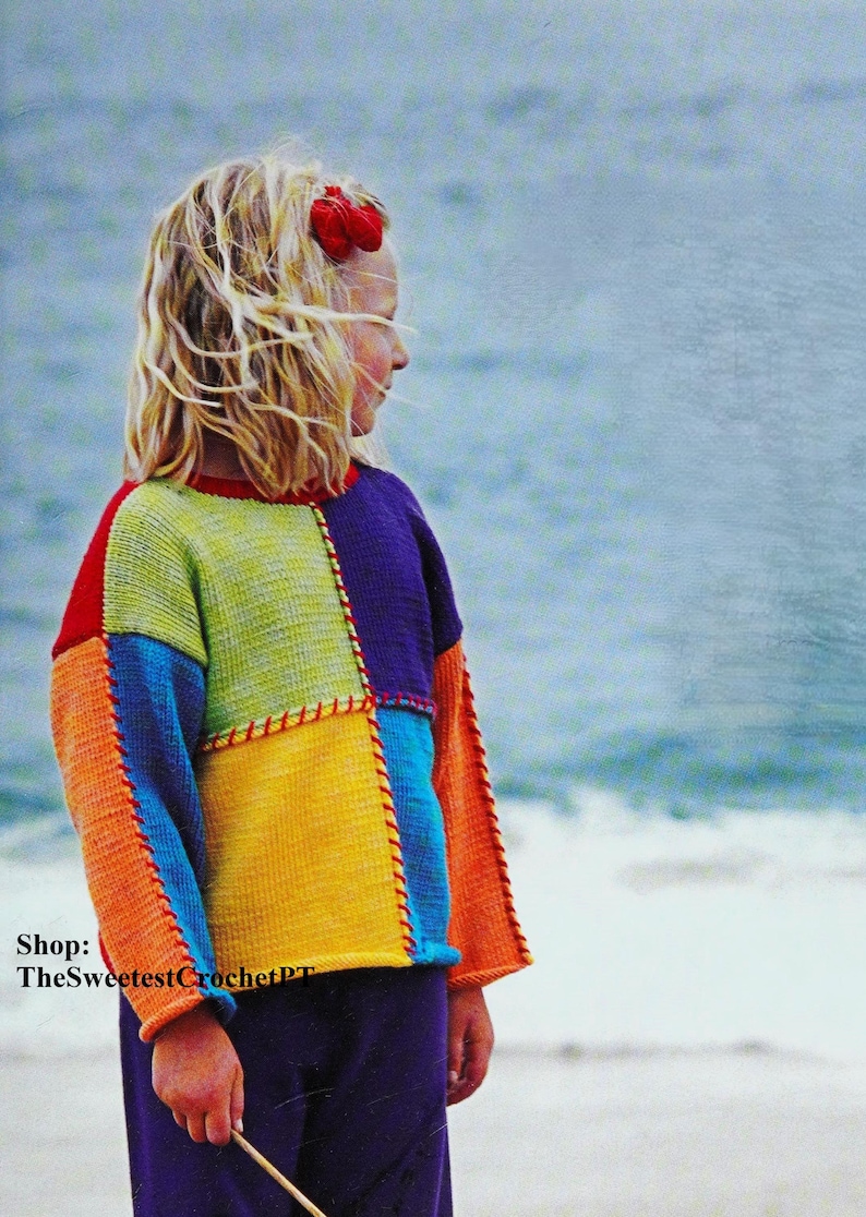 May include: A young person wearing a colourful crocheted sweater with a patchwork design. The sweater is made of different coloured squares of yarn, including orange, yellow, green, blue, and purple. The person is standing on a beach with the sea in the background. The text "Shop: TheSweetestCrochetPT" is visible in the bottom left corner of the image.