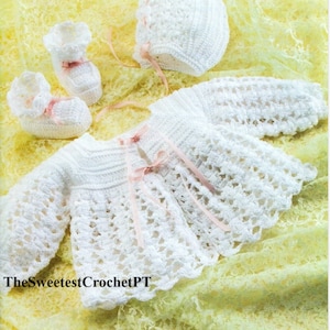 May include: White crocheted baby outfit set including a hat, sweater, and booties. The set is decorated with pink ribbon.