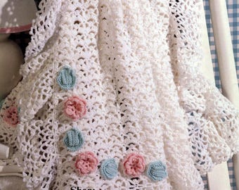May include: A white crocheted baby blanket with a scalloped edge and floral embellishments. The blanket features pink and light blue crocheted flowers. The blanket is draped over a white chair with a blue and white checkered background.