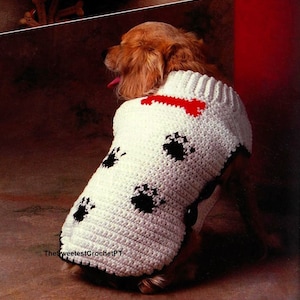 Puede incluir: Un su&eacute;ter de ganchillo blanco para perro con cuello acanalado, un motivo rojo en forma de hueso cerca del escote y patrones de huellas negras en la espalda. La prenda tiene un borde negro en la parte inferior e incluye el texto TheSweetestCrochetPT.